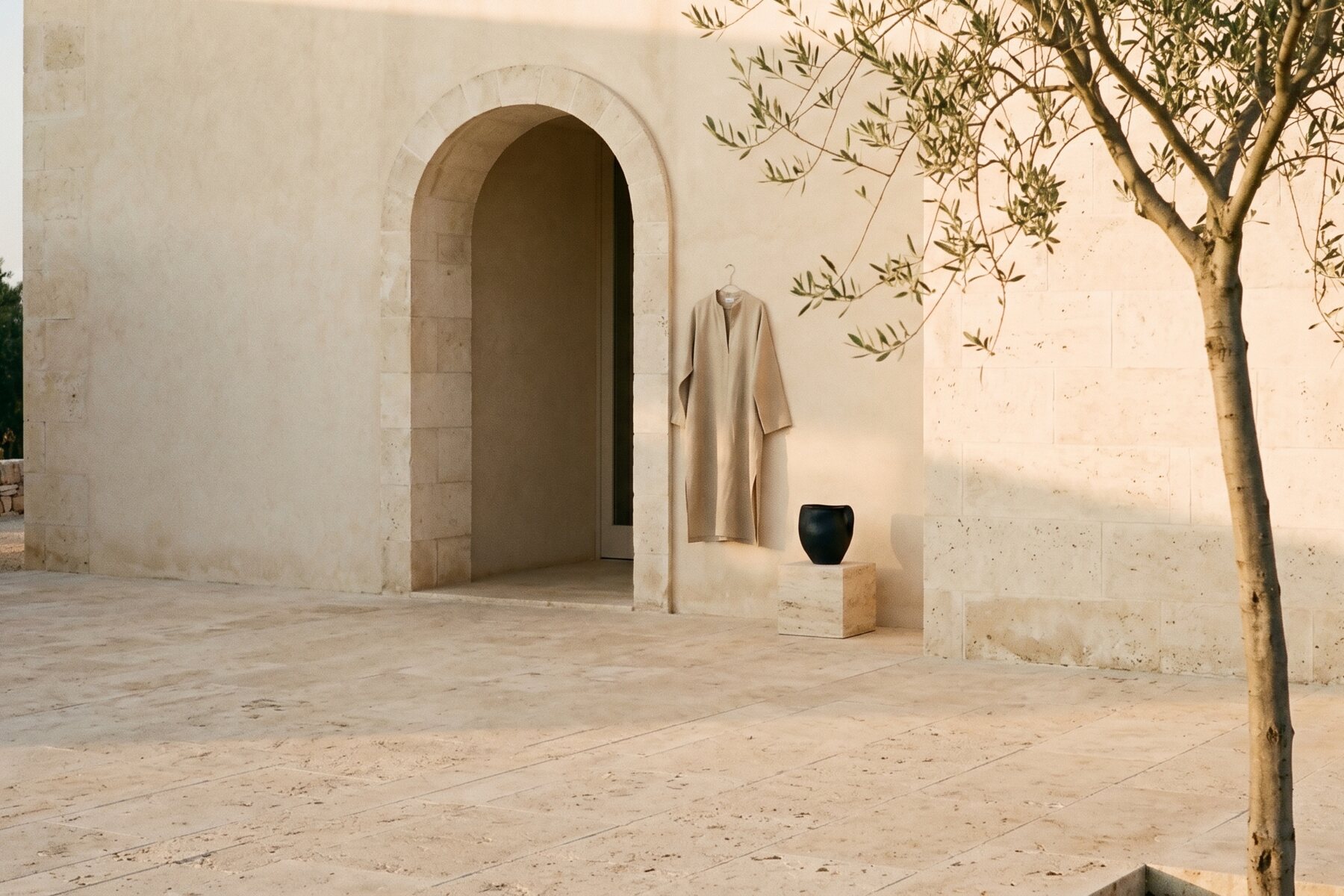 An ARKEN linen garment hanging in a sunlit Mediterranean courtyard, with an olive tree and travertine stone walls