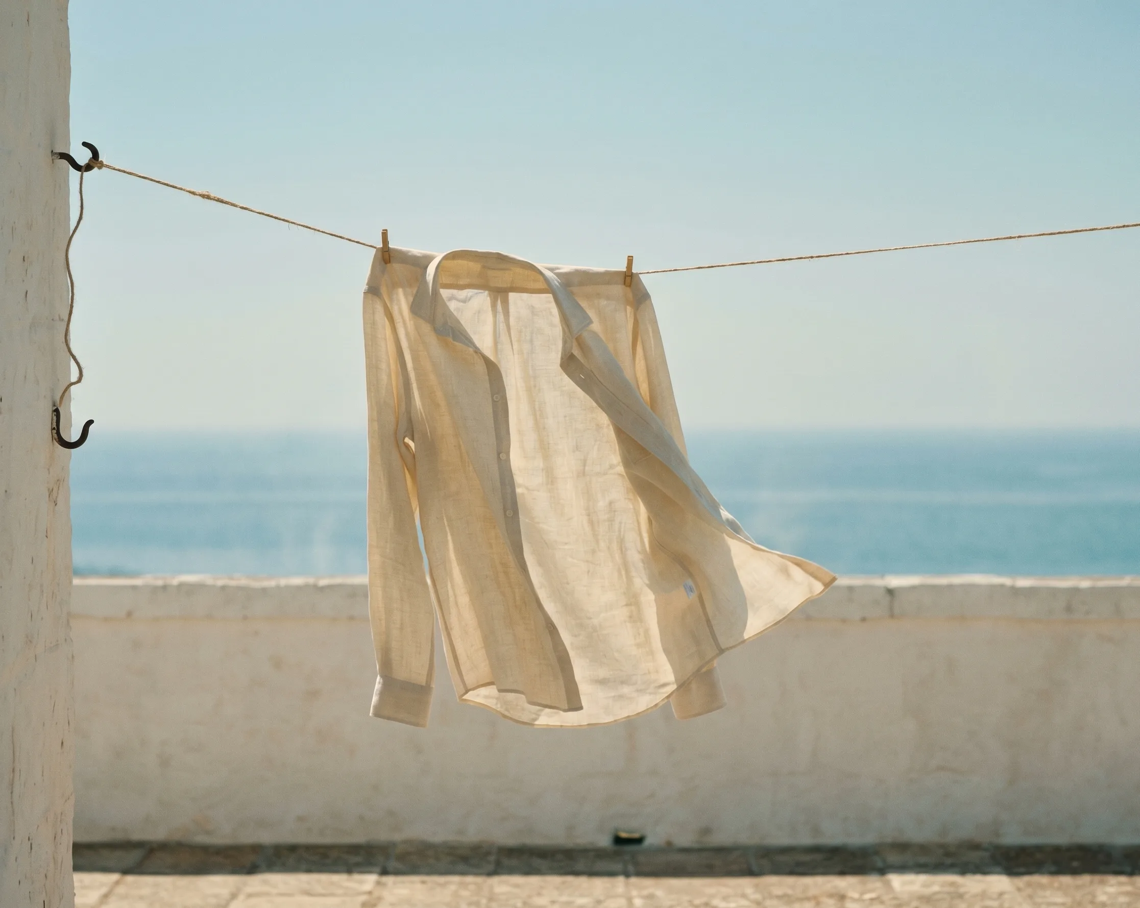 A cream linen shirt on a natural-jute clothesline strung across a low whitewashed parapet, the Adriatic hazy behind, a soft breeze catching one corner of the cloth