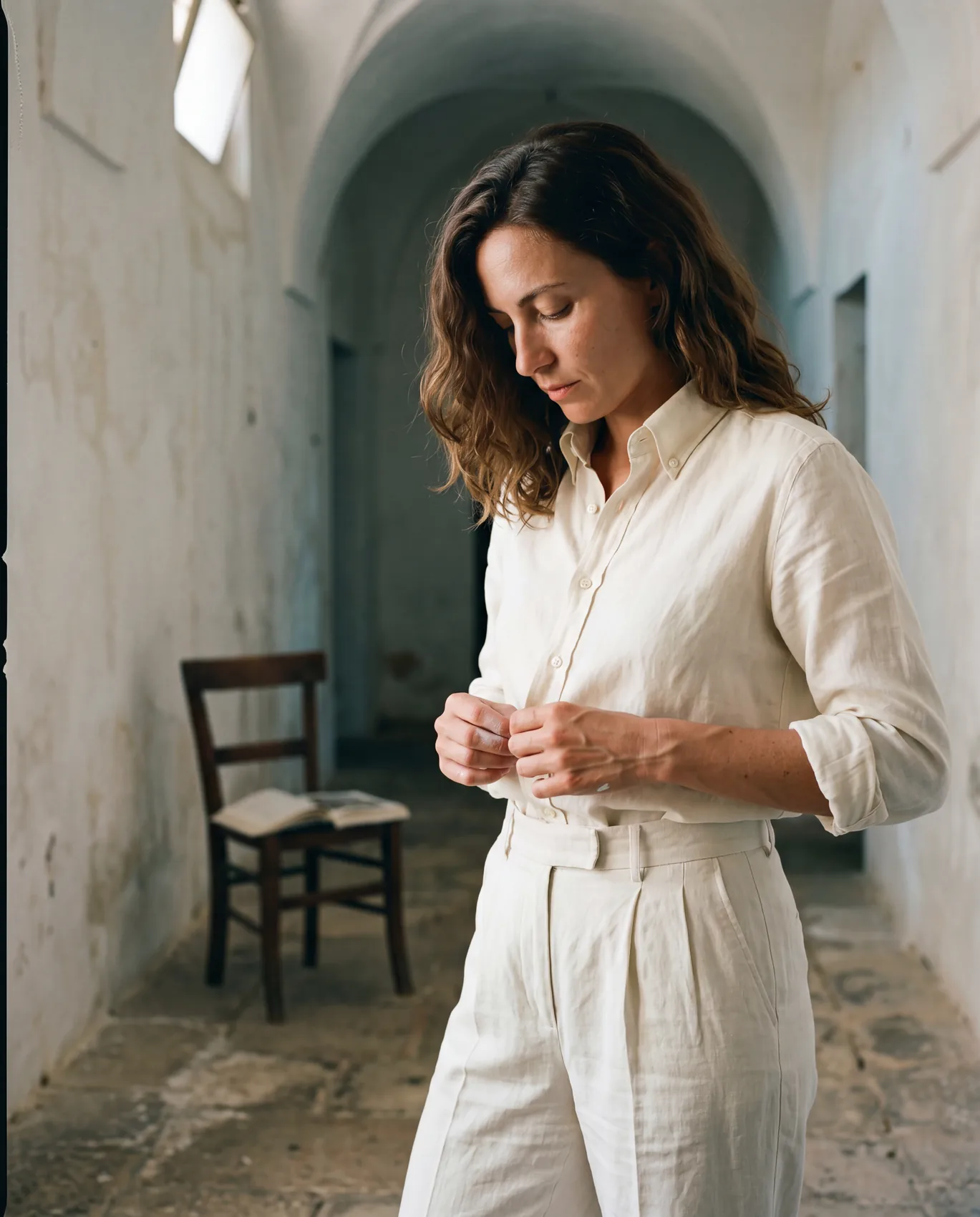 A woman in Bone Mirror trousers and cream Hollow shirt in a whitewashed vaulted hallway, mid-action adjusting a bottom shirt button.