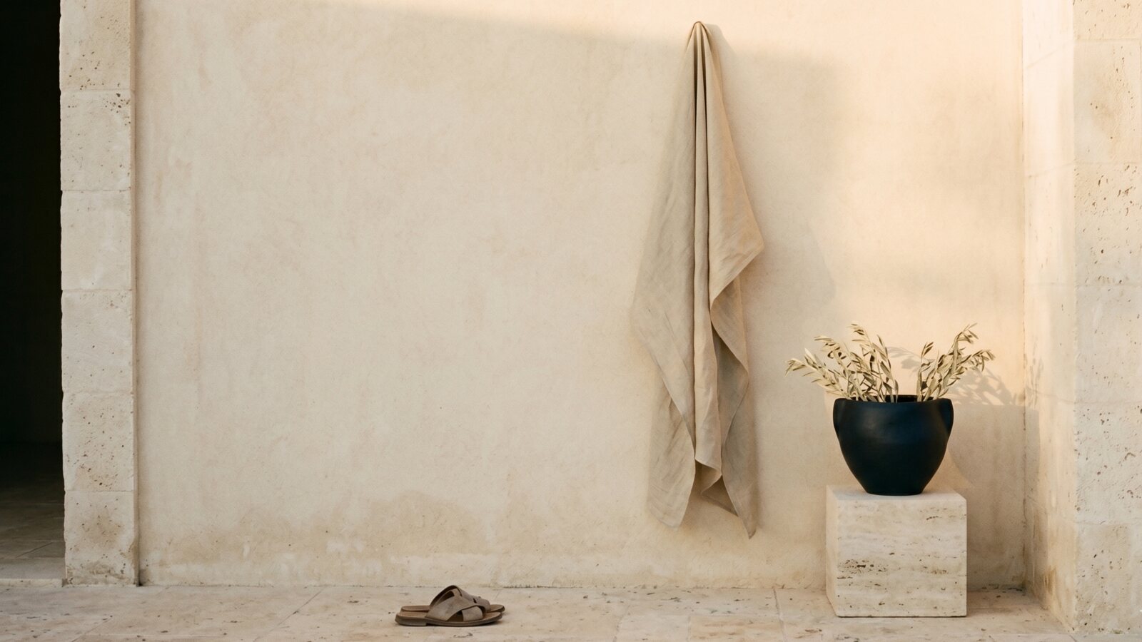 Linen sheet hanging on a warm parchment plaster wall, with leather sandals and olive plant