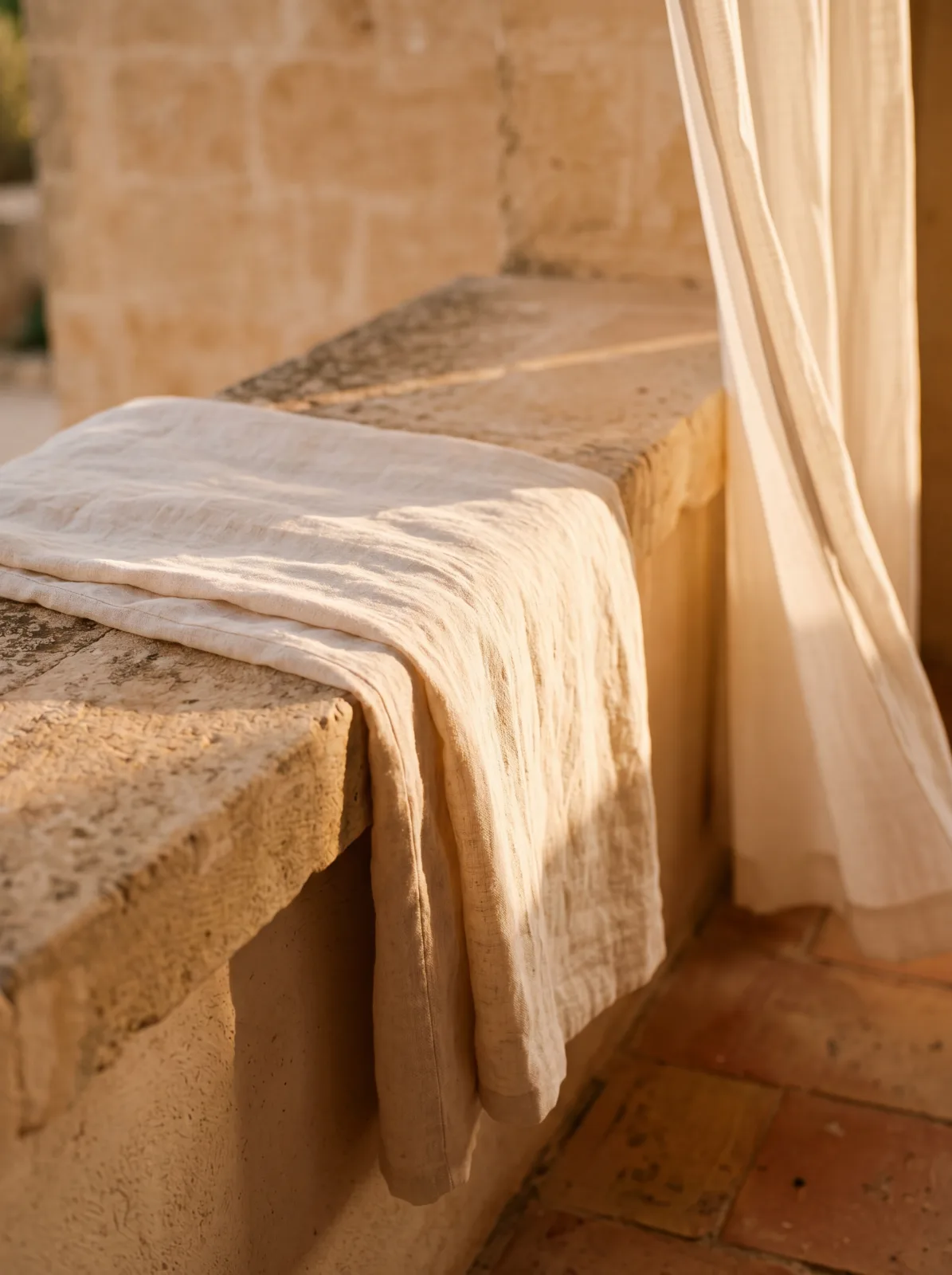 A length of cream linen folded over a worn limestone parapet inside the masseria loggia, sheer ivory curtain at the right edge, warm late-afternoon raking light, terracotta floor below.