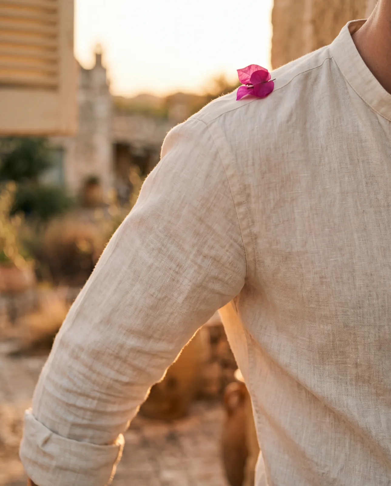 A close-up of a man's cream linen band-collar shirt — sleeve and shoulder both in frame, a single magenta bougainvillea petal fallen at the shoulder seam, the wood shutter and masseria architecture soft-focus behind in the late afternoon light.