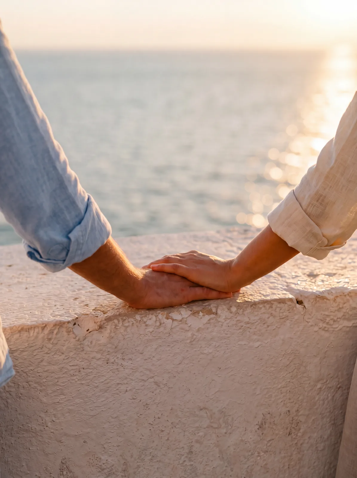 Two hands meeting on the whitewashed stone parapet of the Adriatic terrace at golden hour — the larger left hand emerging from a pale-blue linen sleeve, the smaller right hand from a cream linen sleeve, both rolled to mid-forearm. The soft-focus sea horizon behind, sun catching the water at upper right.