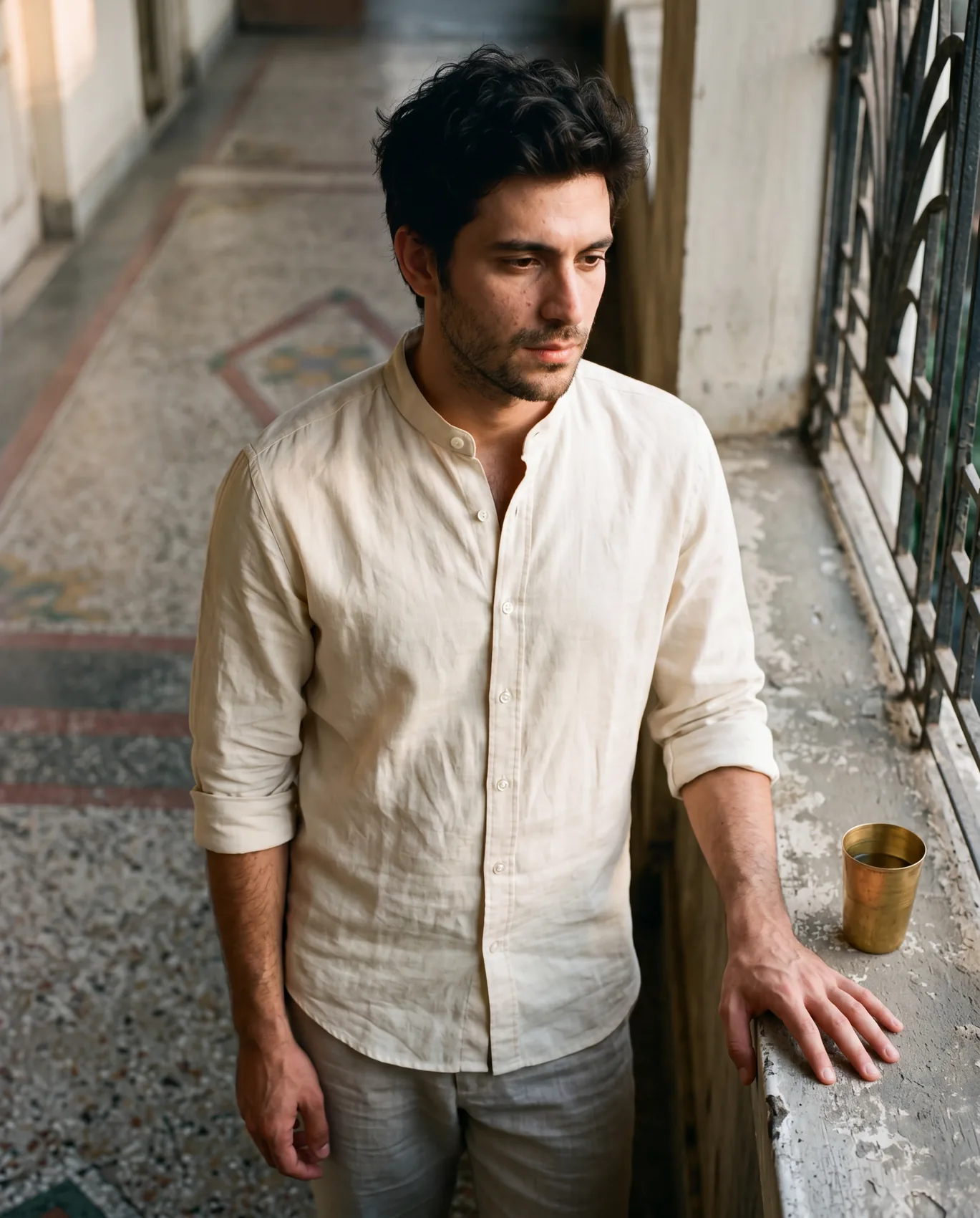 A man in a Parchment Bare shirt at a 1930s Bombay art-deco verandah, three-quarter front pose with the 3cm standing band collar clearly visible at the throat. A brass tumbler rests on the parapet wall beside him.