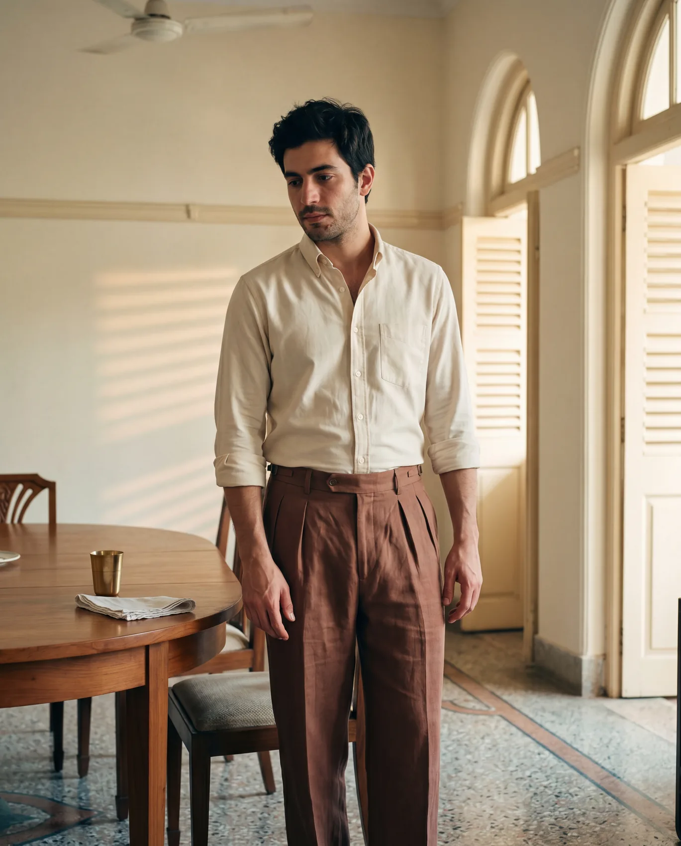A man in a White Hollow shirt and Tobacco Column Wide trousers in a 1930s Bombay art-deco dining room, late afternoon louvre-shadow bands across the wall behind him. A brass tumbler and folded napkin rest on a round teak table beside him.