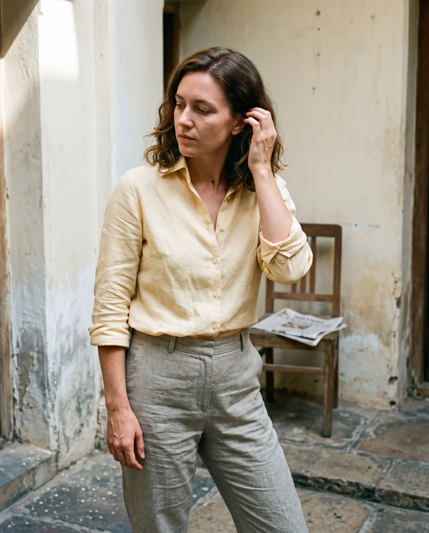 A woman in a Butter Cutaway shirt and stone Column trousers in a Pondicherry stone courtyard, late morning cool light. Caught mid-gesture tucking a strand of hair behind her left ear.