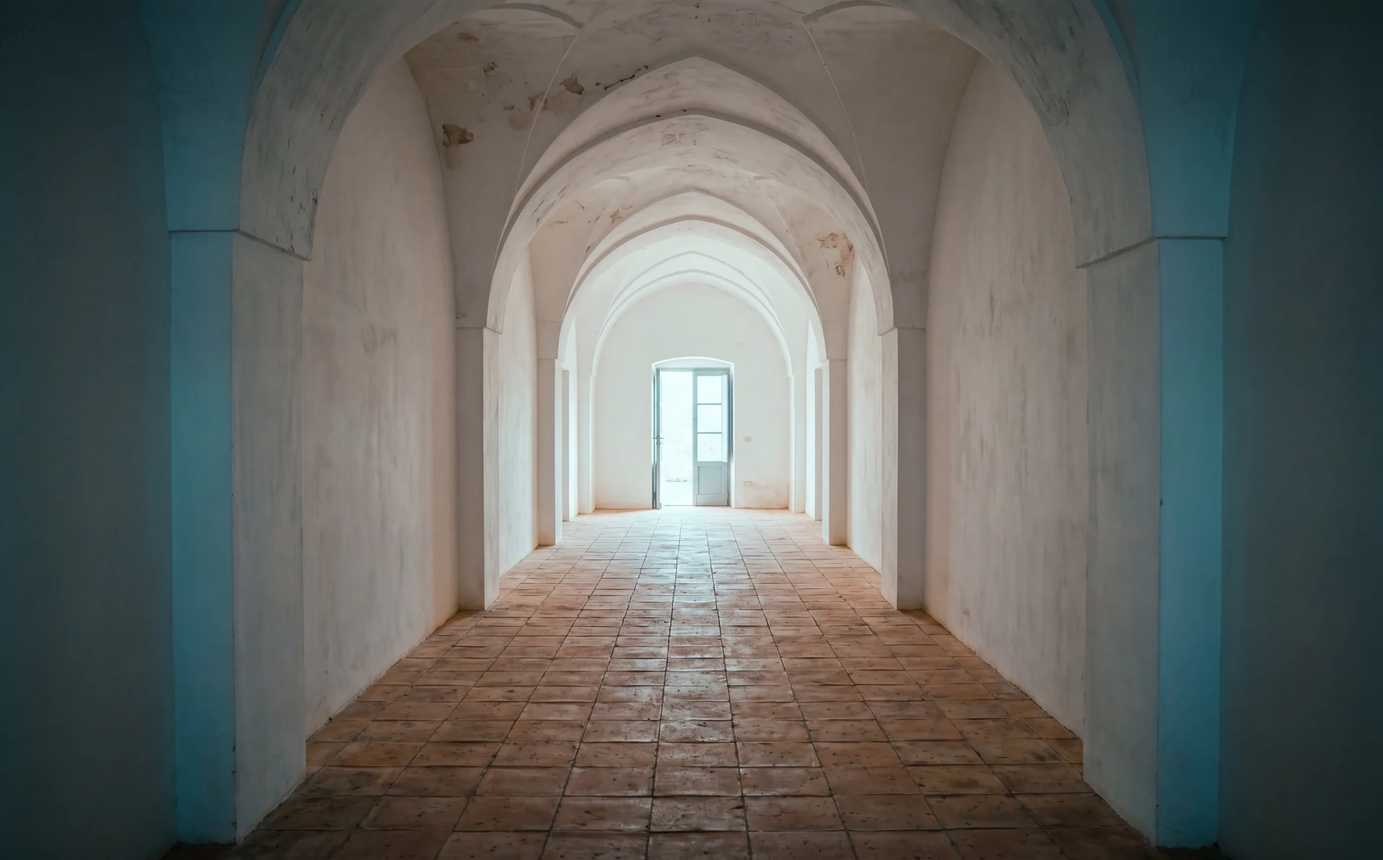 A long whitewashed stone corridor at dawn, vaulted ceiling, terracotta floor, cool light at the far doorway.