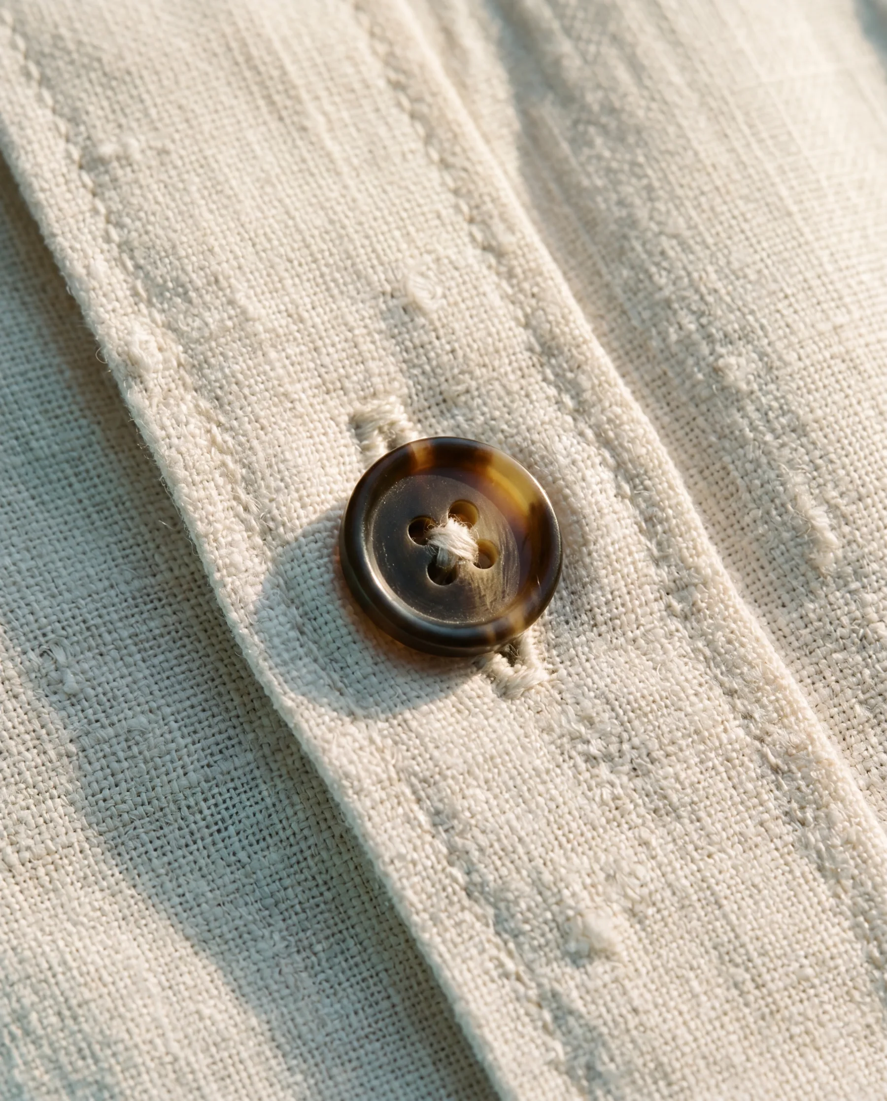 Macro close-up of a horn button on a linen buttonhole, visible weave and irregular slubs in the cloth.