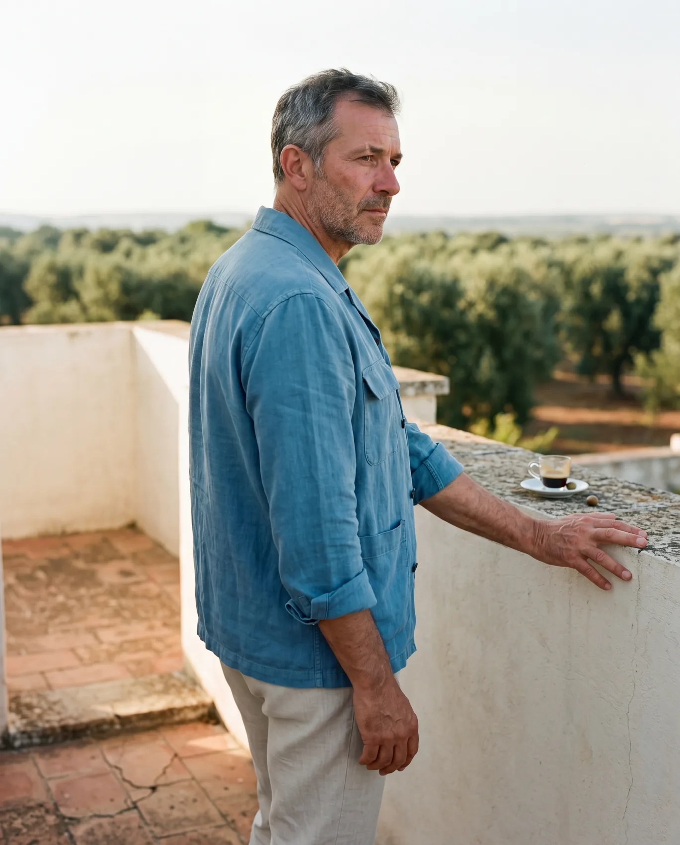 An older man in a Cerulean Frame overshirt buttoned closed, on a masseria rooftop terrace at 5pm. He looks out over olive groves stretching to the horizon, right hand resting on the parapet beside a small espresso cup.