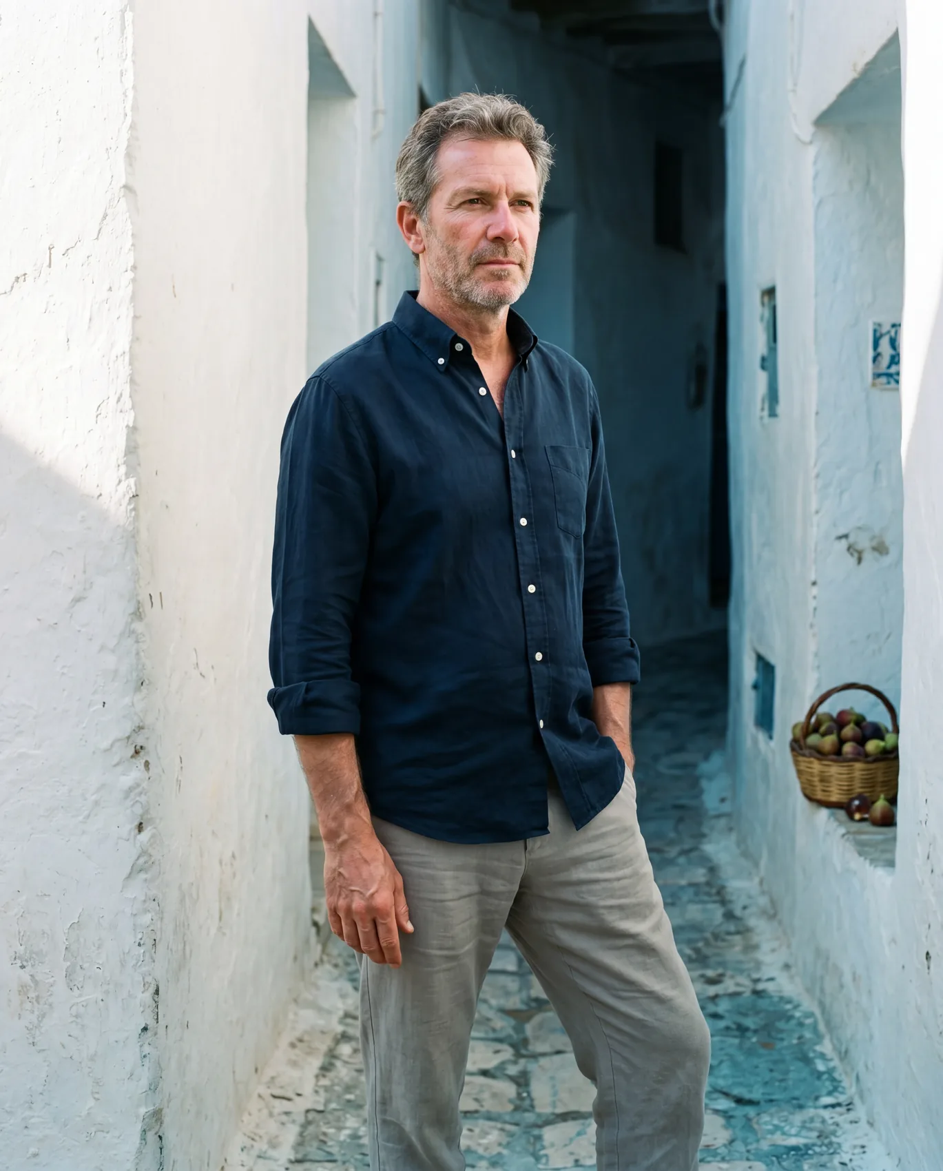 An older man in an Ink Hollow shirt and stone Column trousers in a narrow Tunisian whitewashed alleyway at mid-morning, hard sun on one wall and deep cool shadow on the other. Right hand tucked into the trouser pocket, eyes directed past the camera.