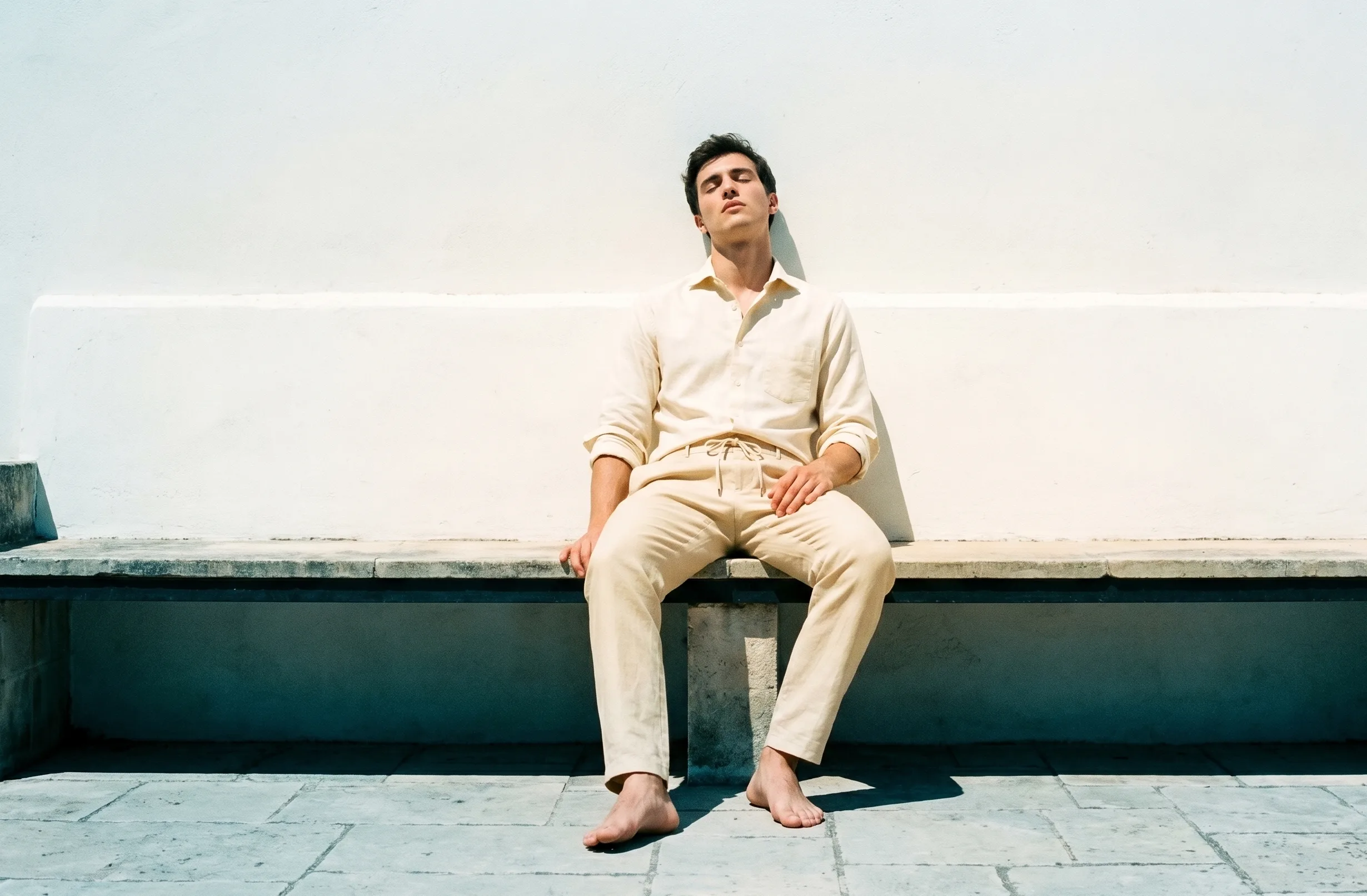 A young man seated on a stone bench against a whitewashed masseria wall at midday, leaning back, eyes closed. Cream Hollow shirt, parchment Ease drawstring trousers, no shoes.