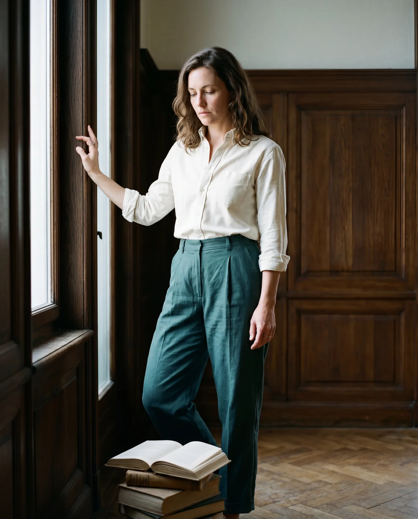A woman in cream Hollow shirt and Seaweed Mirror trousers in a dark oak-panelled library, hand on a tall window frame, eyes directed down at a stack of vintage hardback books on the floor.