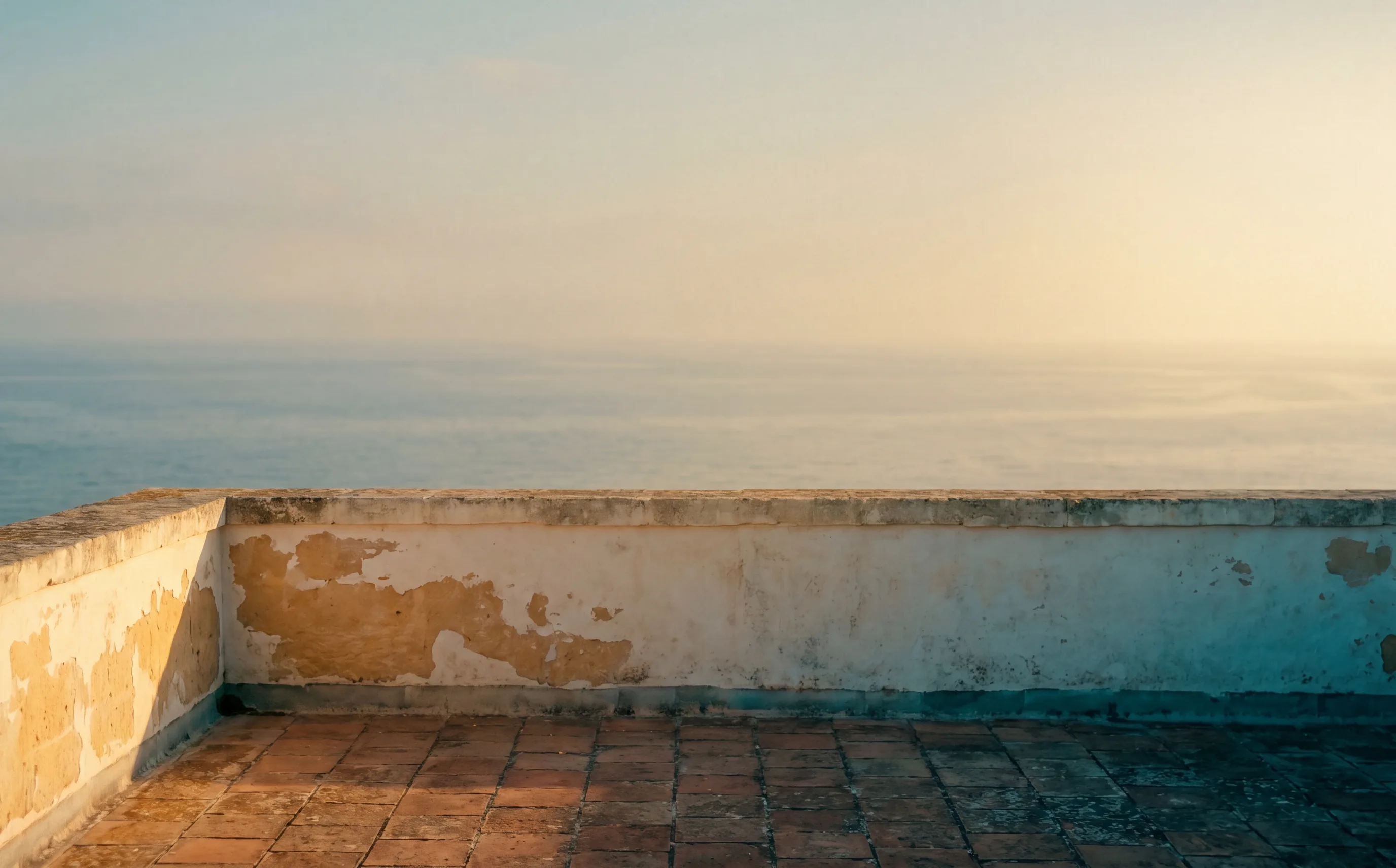 An empty stone terrace at golden hour, low parapet wall, the sea hazy behind.
