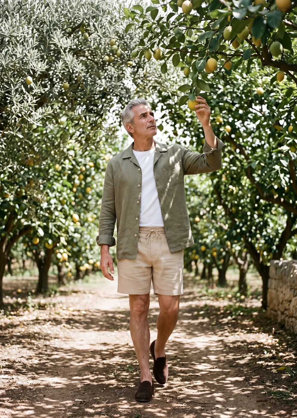 A man in a sage-olive linen overshirt over a pale chambray shirt and stone wide-leg pleated linen trousers, leaning against a weathered Apulian villa wall in soft afternoon light
