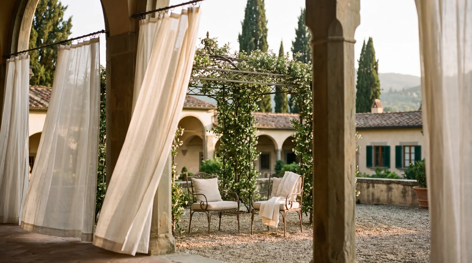 A Tuscan villa loggia in late afternoon — wrought-iron pergola with climbing jasmine, vintage chairs, sheer curtains in foreground, cypress trees beyond.